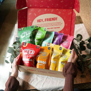 Open box displaying a variety of Hey! Chips products including signature collection broccoli, okra, mango, banana, and more.