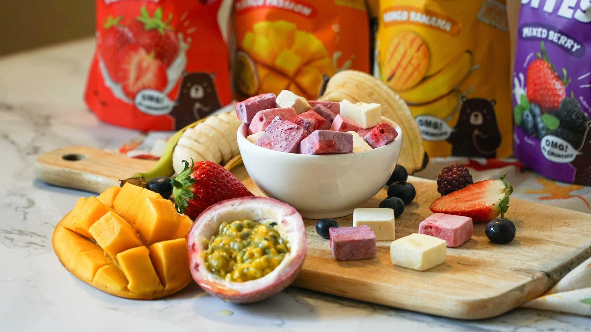 Assorted freeze-dried fruit cubes in a white bowl surrounded by fresh mango, strawberry, banana, passion fruit, blueberries, and blackberries, with Hey! Chips fruit snack packaging in the background.