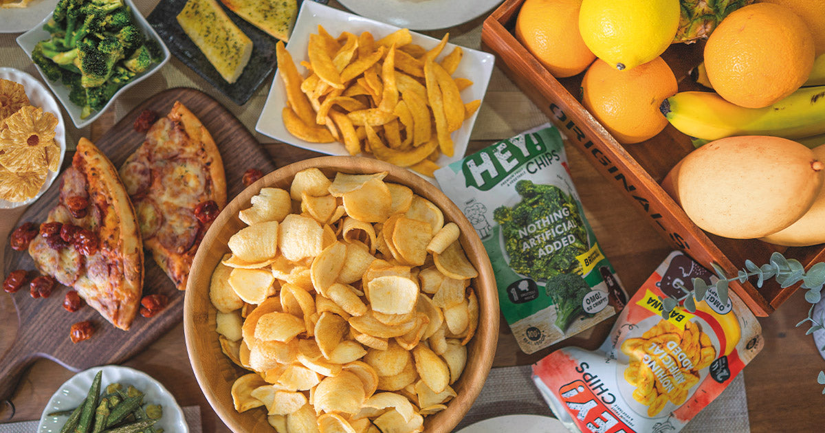 A table with various snacks, including a large bowl of potato chips, a bag of Hey! Chips, pizza, vegetables, and fruit