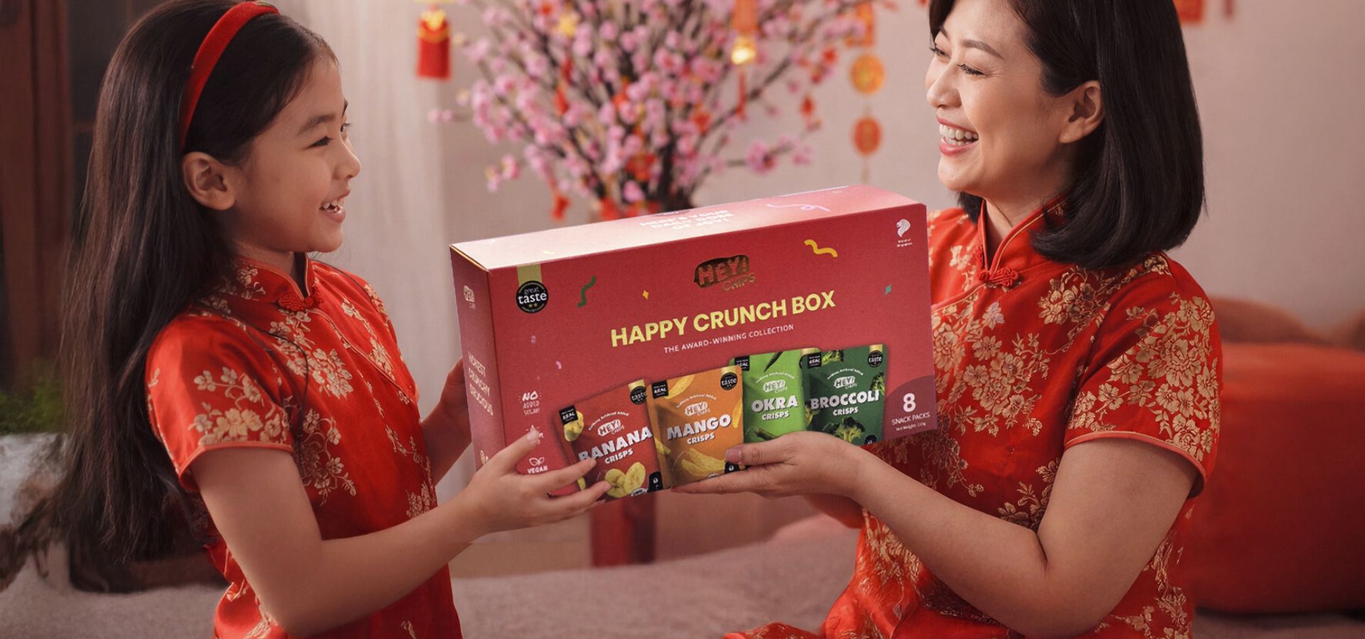 Two women in traditional red outfits opening a Hey! Chips 'Happy Crunch Box' in a festive setting.