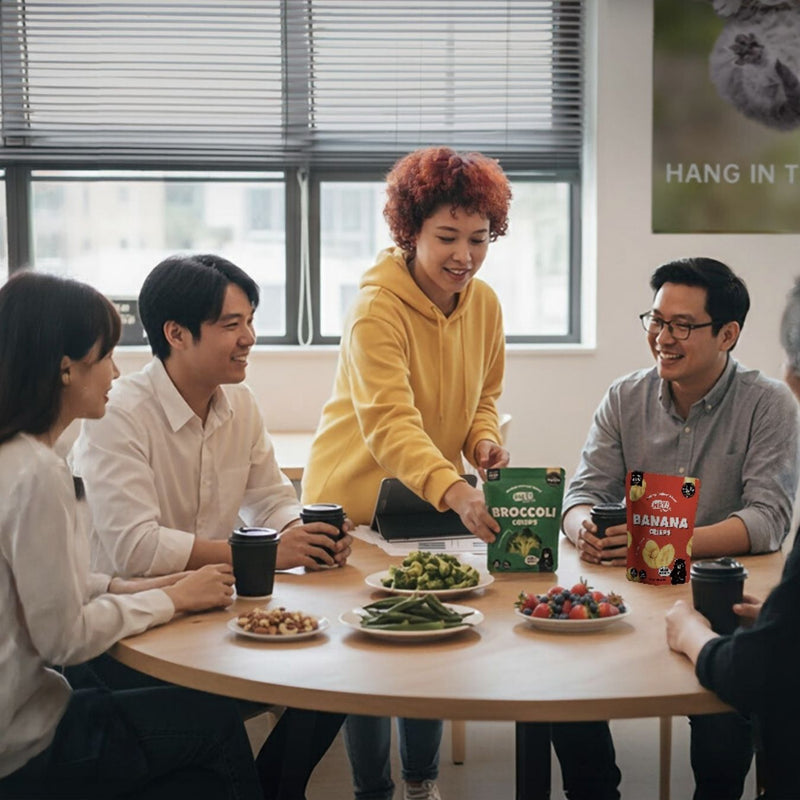 Group of people sitting around a table with snacks and drinks, one person is holding a Hey! Chips broccoli.